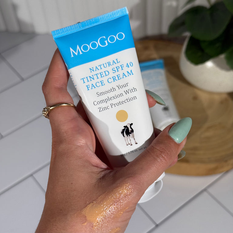Hand holding a tube of MooGoo Natural Tinted SPF 40 Face Cream with a swatch on skin, with a tile background featuring a plant and wooden plate.