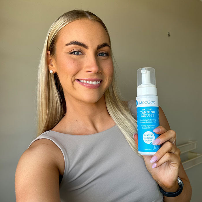 MooGoo Natural Tanning Mousse held in hand by smiling woman, lightweight self tan mousse for streak-free golden glow, nourishing and buildable.