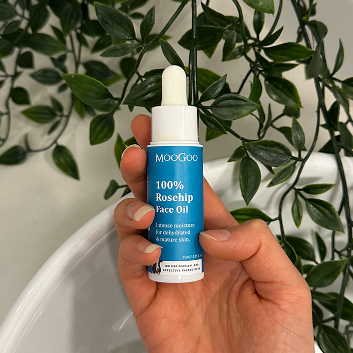 A hand holding MooGoo 100% Rosehip Face Oil bottle in leafy background, gives intense moisture for dehydrated & mature skin.