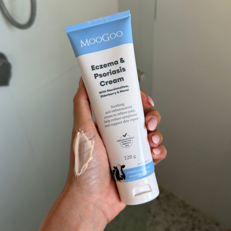 A hand holds a squeeze tube of MooGoo Eczema & Psoriasis Cream, showing a smear of the beige cream on the back of the hand against a blurred bathroom background.