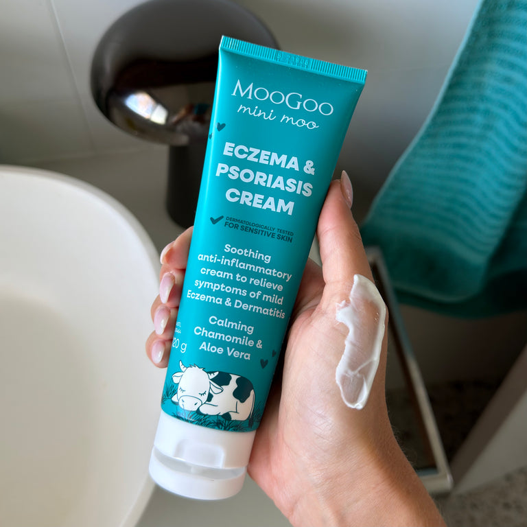 A hand holding a tube of MooGoo Mini Moo Eczema & Psoriasis Cream, with white cream smeared on the back of the hand, set against a bathroom background.