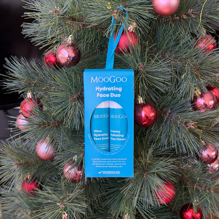 MooGoo Hydrating Face Duo skincare set in its blue hanging box with one mini creamy hydrating face cleanser and one ultra hydrating face cream, displayed on a Christmas tree with green needles and red ornaments.