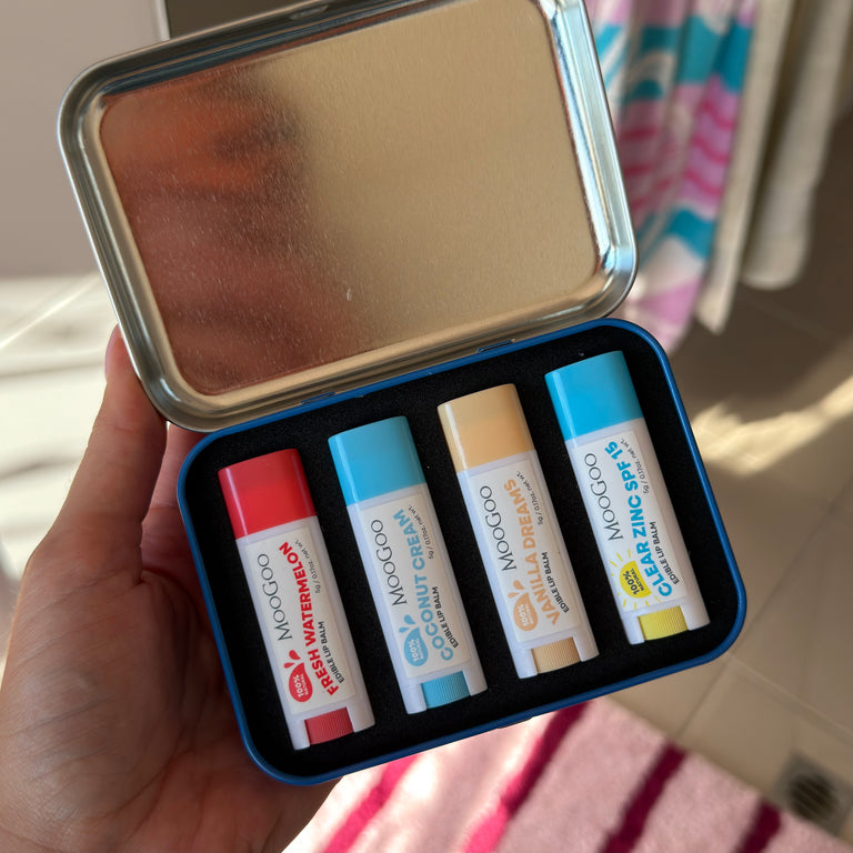A hand holds an open silver metal tin containing four MooGoo lip balm tubes in different colors and flavours: red fresh watermelon, light blue coconut cream, beige vanilla dream, and dark blue clear zinc spf15. The background shows a domestic bathroom setting with tiled floors and a shower curtain.