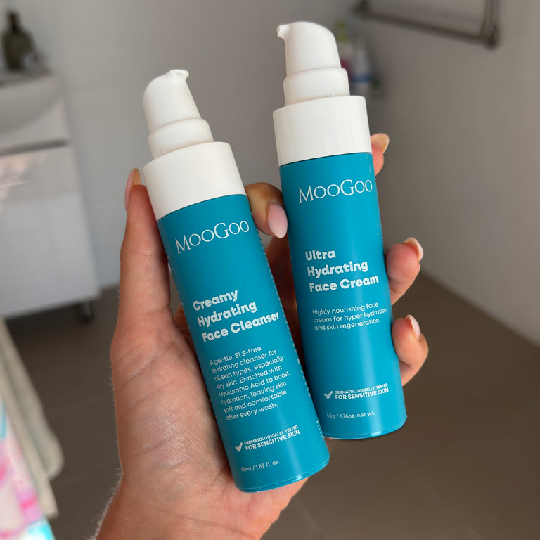 A hand holding two MooGoo skincare bottles, one mini creamy hydrating face cleanser and one ultra hydrating face cream, in a bathroom setting. 