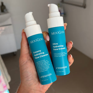 A hand holding two MooGoo skincare bottles, one mini creamy hydrating face cleanser and one ultra hydrating face cream, in a bathroom setting. 