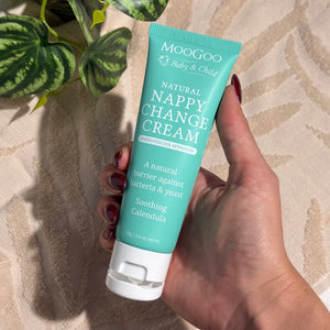 Hand holding a tube of MooGoo Baby & Child Natural Nappy Change Cream on a beige towel background with a green plant, paediatrician approved, calms and protects baby's skin against nappy rash.