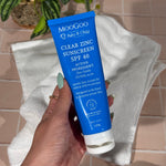 MooGoo Baby Clear Zinc Sunscreen SPF 40 tube held by a hand with a towel and plant in the background against pink tiles.