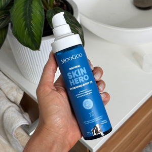 Hand holding a blue bottle of MooGoo Skin Hero Body Oil with a plant in the background on a bathroom sink with towel in the corner. With 6 hero nourishing oils to instantly replenish, soften and hydrate dry skin. Lightweight, Fast-Absorbing & Ideal for Pregnancy & Postpartum Skincare
