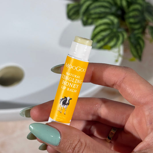 Hand holding a yellow tube of MooGoo Natural Tingling Honey Lip Balm with a plant and bathroom sink in the background.