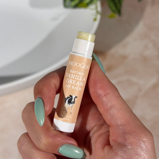 Hand holding a beige tube of MooGoo Natural Vanilla Dream Lip Balm with a plant and bathroom sink in the background.