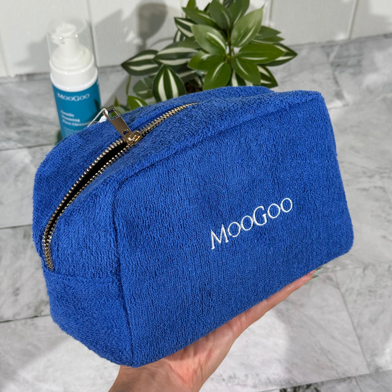Blue MooGoo Terry Towelling Cosmetics Travel Bag with a gold zipper held by a hand with a tiled background, with a plant and MooGoo Foaming Cleanser.