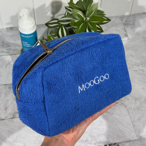 Blue MooGoo Terry Towelling Cosmetics Travel Bag with a gold zipper held by a hand with a tiled background, with a plant and MooGoo Foaming Cleanser.