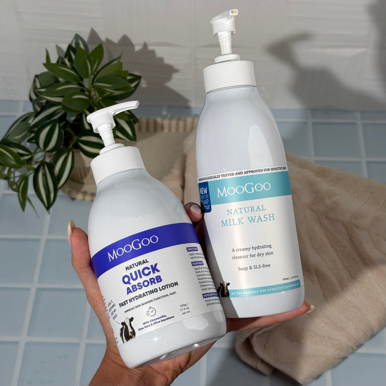 Hand holding bottles of MooGoo Milk Wash and Fast Hydrating Lotion with cream towel, blue tiles and greenery in the background, Creamy Hydrating Cleanser and Lightweight Lotion To Replenish Dry, Sensitive Skin