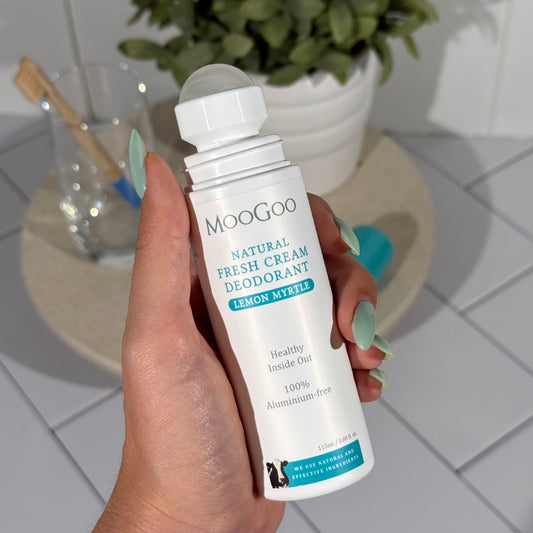 MooGoo Natural Lemon Myrtle Fresh Cream Deodorant 115ml held by a hand with a tiled background and a plant and toothbrush on a tray.