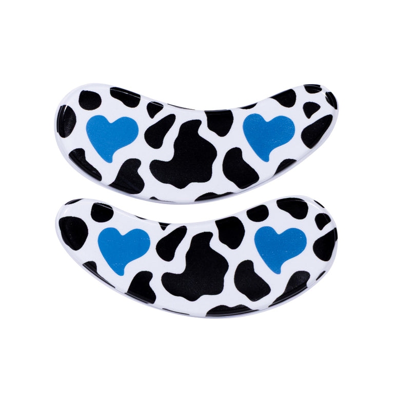 Two curved reusable eye mask patches with a black and white cow print pattern and two blue heart designs, shown on a plain white background. To lock in moisture and hydration from skincare and eye serums. 