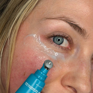 Close-up of a woman's face with a blue skincare rollerball tube being used to apply MooGoo 3-in-1 Peptide Eye Serum under her eye. Made with Vitamin C, Copper Tri-Peptide, Argireline Amplified and Vitamin K to brighten dark circles, reduce fine lines and wrinkles, and diminish under eye puffiness.