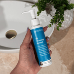 Hand holding MooGoo Oil Cleansing Method 100ml beside sink, non-foaming cleanser with jojoba oil for clear pores, hydration and gentle deep cleansin