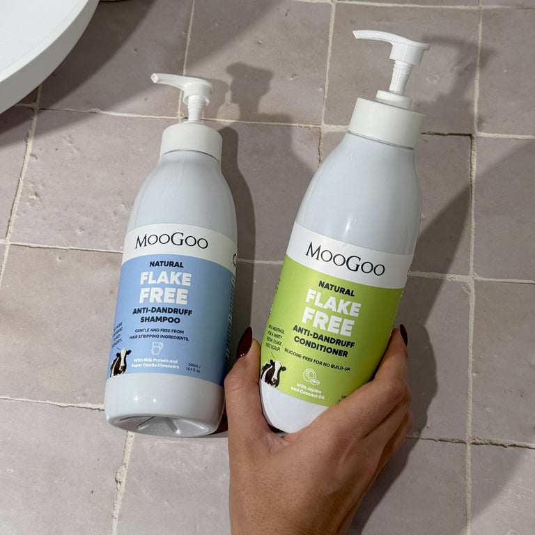 Two bottles of Moogoo Flake Free shampoo and conditioner held against a tiled bathroom wall.