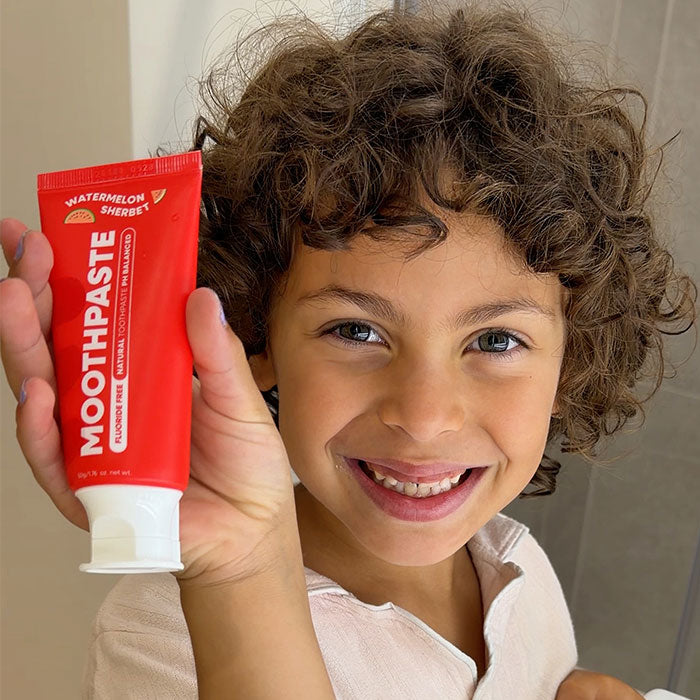 A smiling child with curly brown hair holds up a red tube of Watermelon Kids Moothpaste toothpaste in front of their face. 