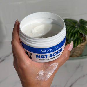 Hand holding a jar of MooGoo Nat Sorb minimalist moisturiser with a plant in the background, pH balanced natural moisturiser for easily irritated and reactive skin
