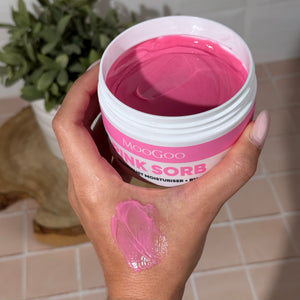Hand holding MooGoo Pink Sorb Cream with pink swatch on hand and greenery and pink tiles in the background. 