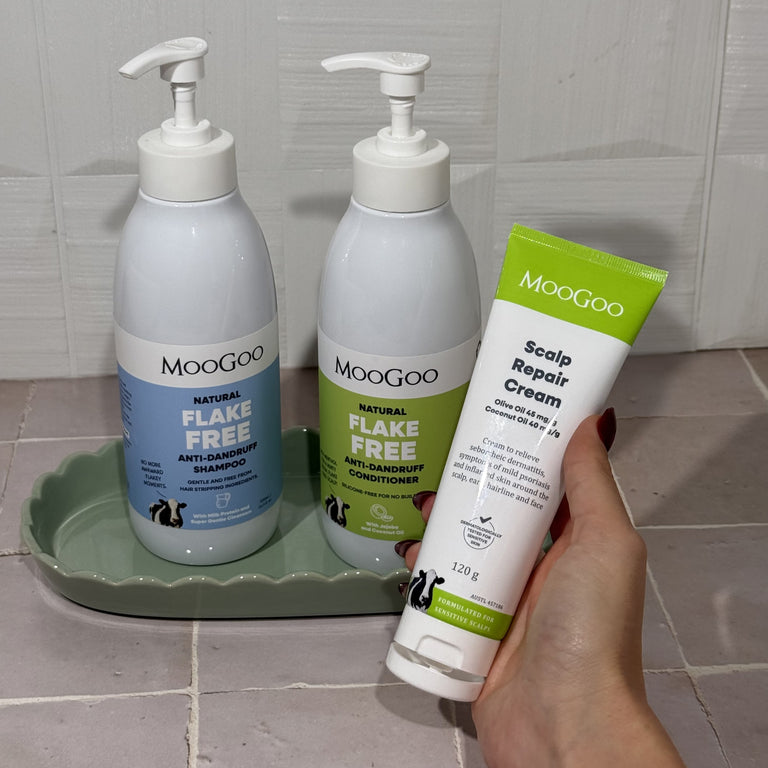 A hand holding MooGoo Flake Free Anti-Dandruff Shampoo, with MooGoo Flake Free Anti-Dandruff Conditioner and MooGoo Scalp Reapir Cream on a bathroom bench, next to a sink in the background, for dandruff and seborrheic dermatitis, gentle on sensitive scalps