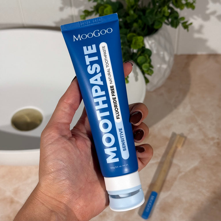 Hand holding a tube of MooGoo Fluoride Free Sensitive Peppermint Whitening toothpaste with a wooden toothbrush in a bathroom setting.