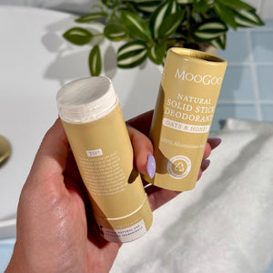 MooGoo 65g Natural Solid Stick Deodorant Oats & Honey held in a hand with the cap off against a bathroom background with a plant. 100% aluminium free, plastic-free natural odour control.