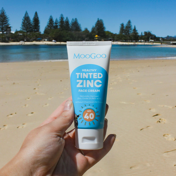 A female hand holding a tube of MooGoo Tinted Zinc Face Cream 50g on a beach with the sand and water in the background.
