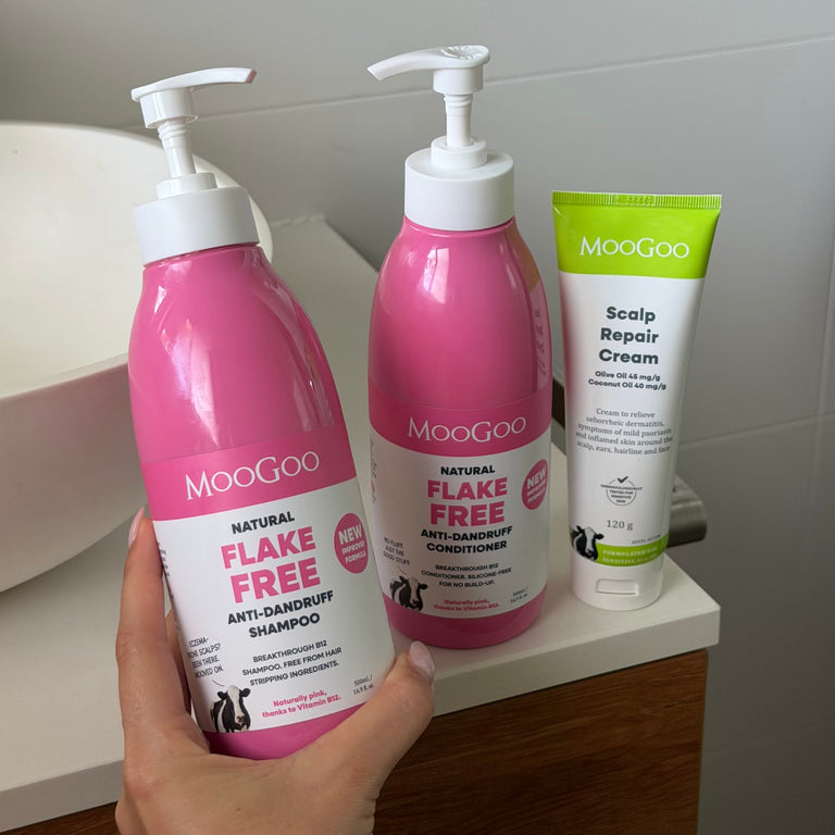 A hand holding MooGoo Pink Flake Free Anti-Dandruff Shampoo, with MooGoo Pink Flake Free Anti-Dandruff Conditioner and MooGoo Scalp Reapir Cream on a bathroom bench, next to a sink in the background, for dandruff and seborrheic dermatitis, gentle on sensitive scalps