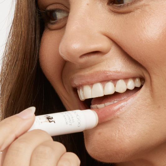 Woman applying MooGoo Natural Coconut Cream Lip Balm on plain white background, nourishing lip care made with edible ingredients for soft hydrated lips, petroleum free, all natural.